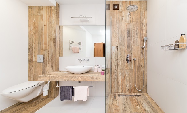 Nowoczesne wnętrze łazienki połączone z toaletą. Prysznic i toaleta wyłożone kafelkami w kolorze drewna, białe ściany i biała podłoga oraz umywalka z dużym lustrem pośrodku